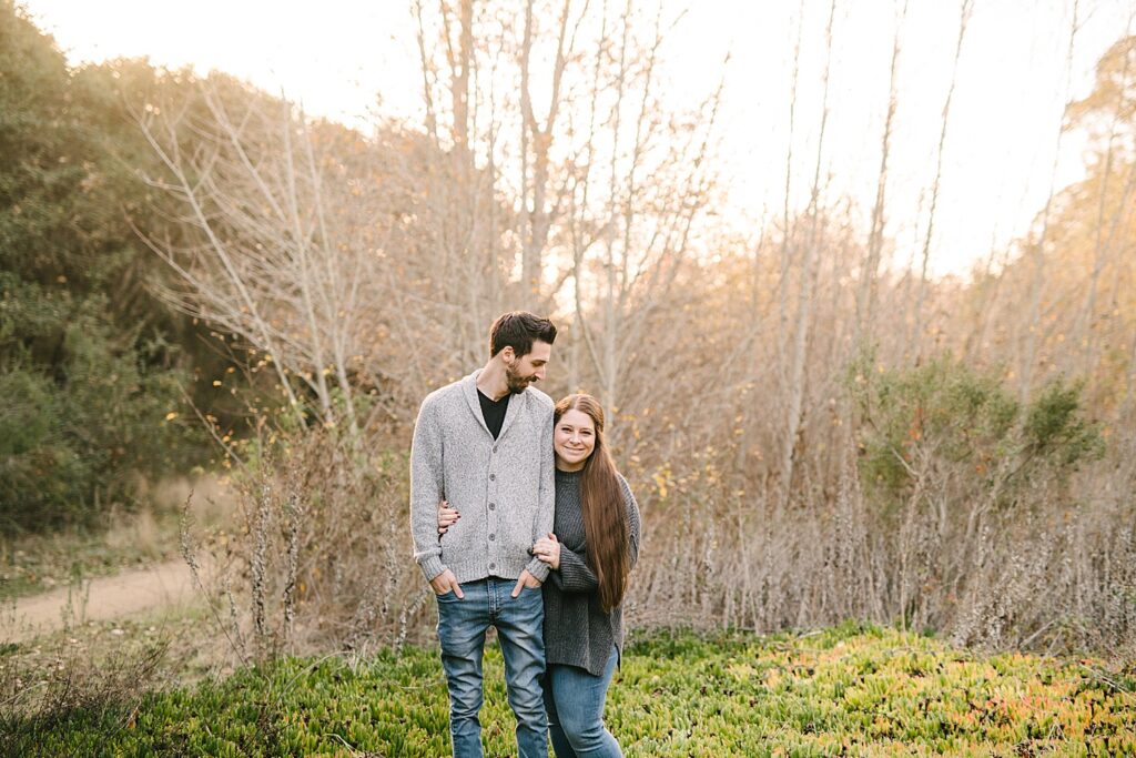 Wife stands slightly behind her husband, peaking around at the camera while he looks down at her on nature trail by Arroyo Grande family photographer Austyn Elizabeth Photography