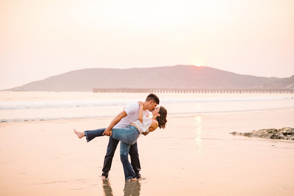 Dipping his bride on the beach with the golden ash sunset during California fire season at Avila Beach engagement session by Avila Beach engagement photographer Austyn Elizabeth Photography