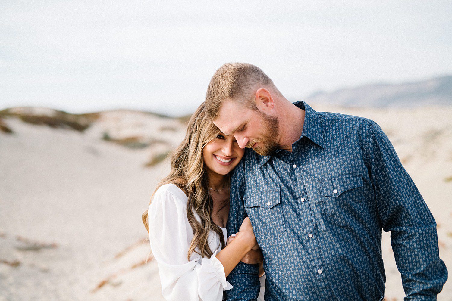 Girl smiles and tucks into guy's neck during engagement session on Pismo Beach Dunes by Pismo Beach Engagement Photographer Austyn Elizabeth Photography