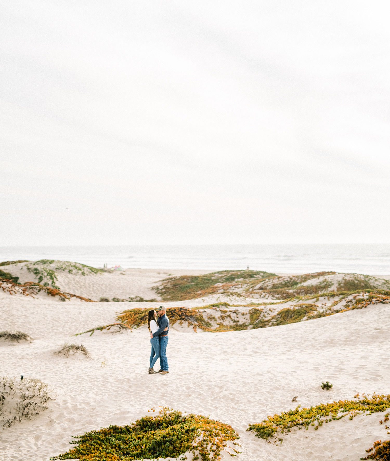 Couple kisses in Pismo Beach dunes with Pacific Ocean in background by Pismo Beach Engagement Photographer Austyn Elizabeth Photography
