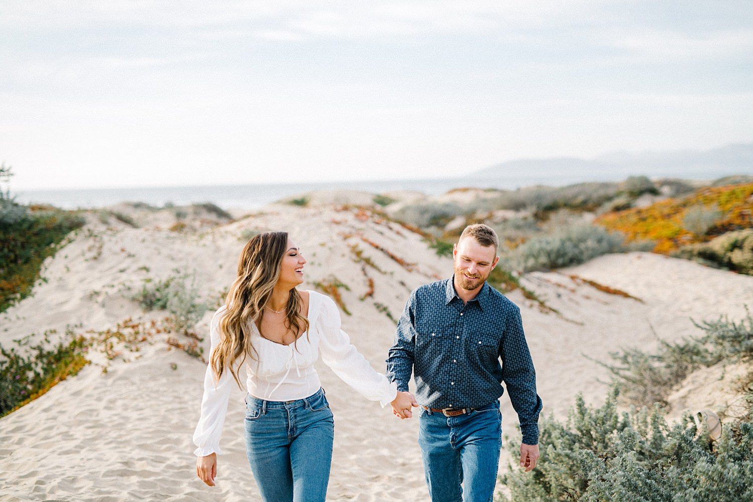 Couple holding hands running through Pismo Beach dunes with Pacific Ocean in background by Pismo Beach Engagement Photographer Austyn Elizabeth Photography