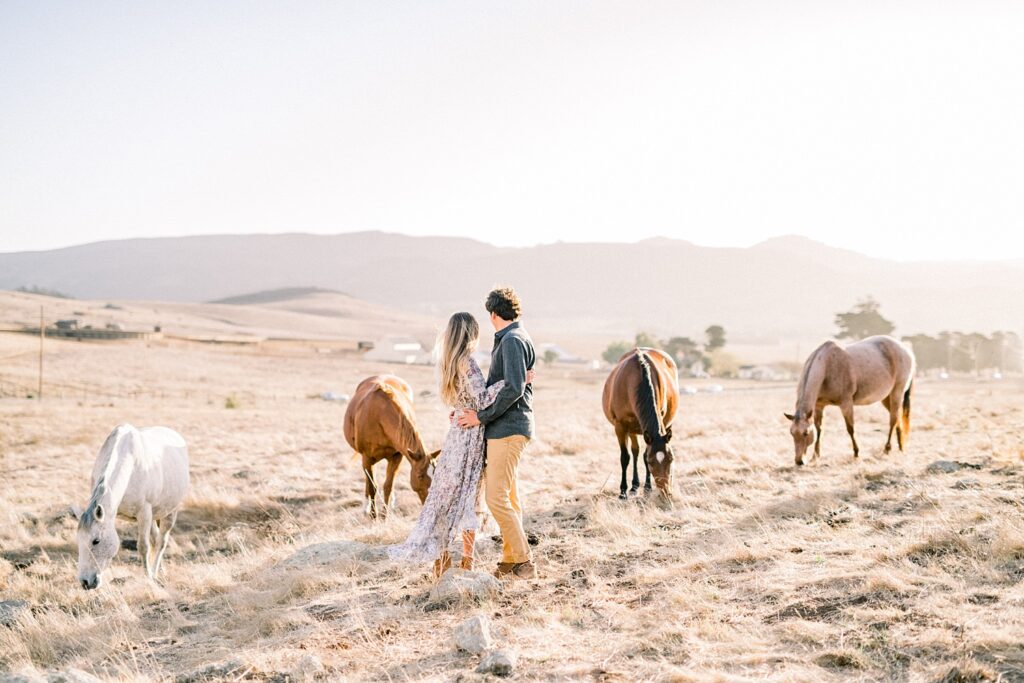 Couple standing surrounded by horses in field and anthropology engagement dress by San Luis Obispo Wedding Photographer Austyn Elizabeth Photography