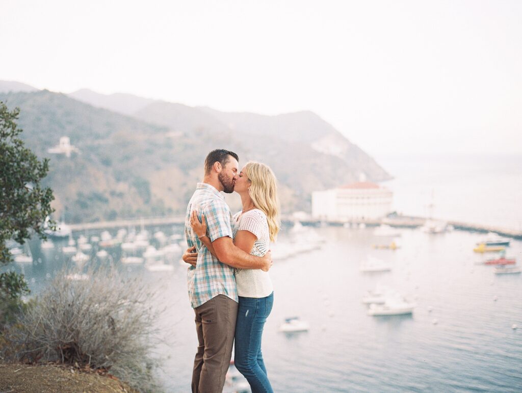 Couple preparing for their Della Terra Mountain Chateau Wedding in the Rocky Mountains, hold and kiss while ontop of cliff overlooking Catalina Casino by Avalon Wedding Photographer Austyn Elizabeth Photography
