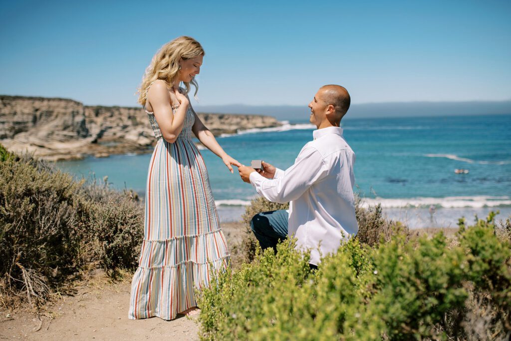 holding her hand and he on one knee at surprise proposal at Montana De Oro's Spooners Cove by Pismo beach engagement photographer austyn elizabeth photography