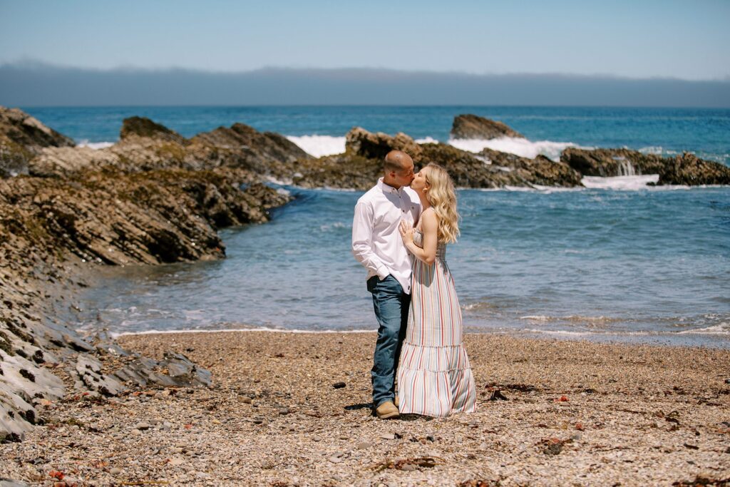 Blue ocean and blue skies while couple stands on beach and kisses at surprise proposal at Montana De Oro's Spooners Cove by Pismo beach engagement photographer austyn elizabeth photography