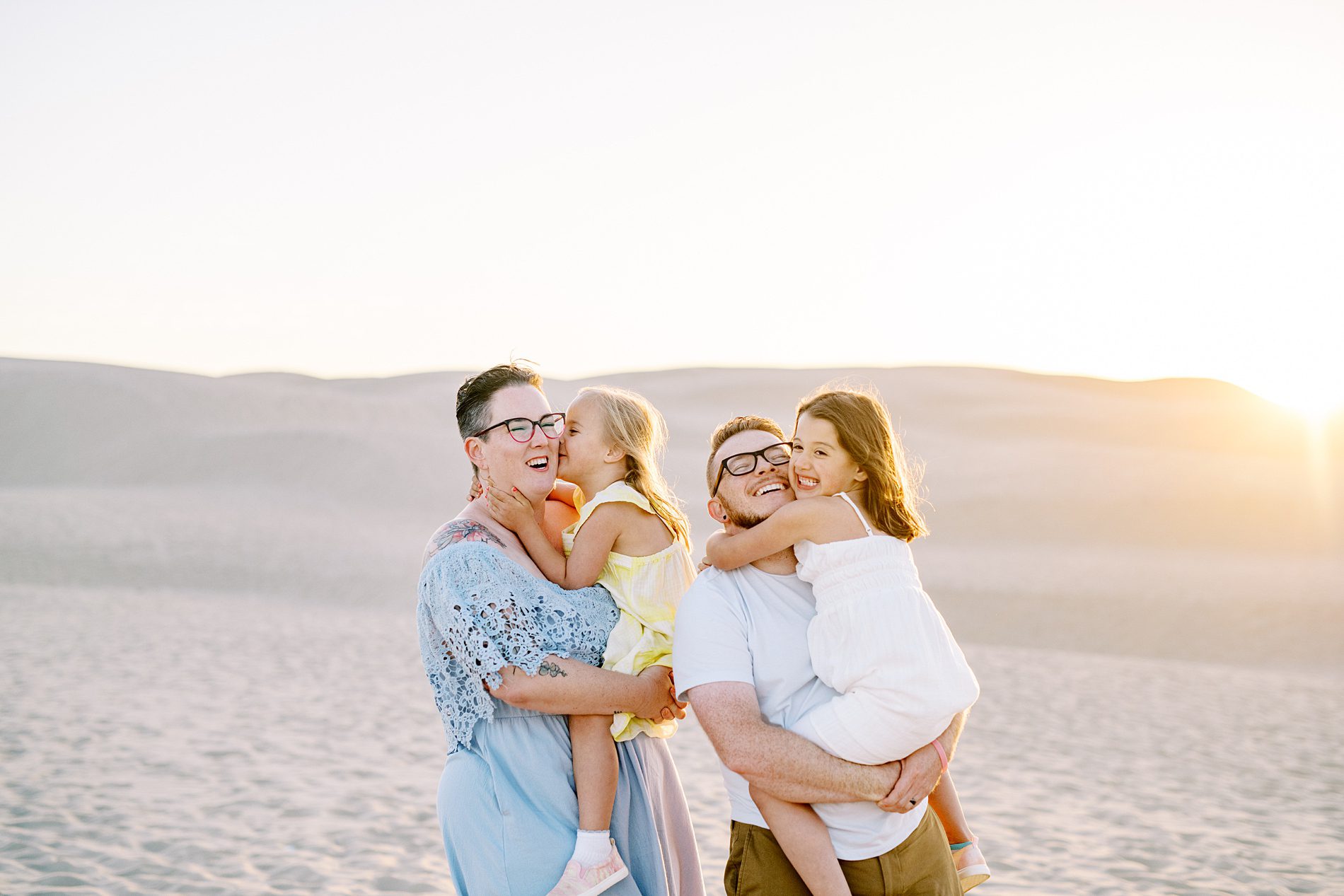 Mom and dad hold their two girls with the rolling big white dunes in the background at sunset at the Oceano Dunes by Pismo Beach Family Photographer Austyn Elizabeth Photography
