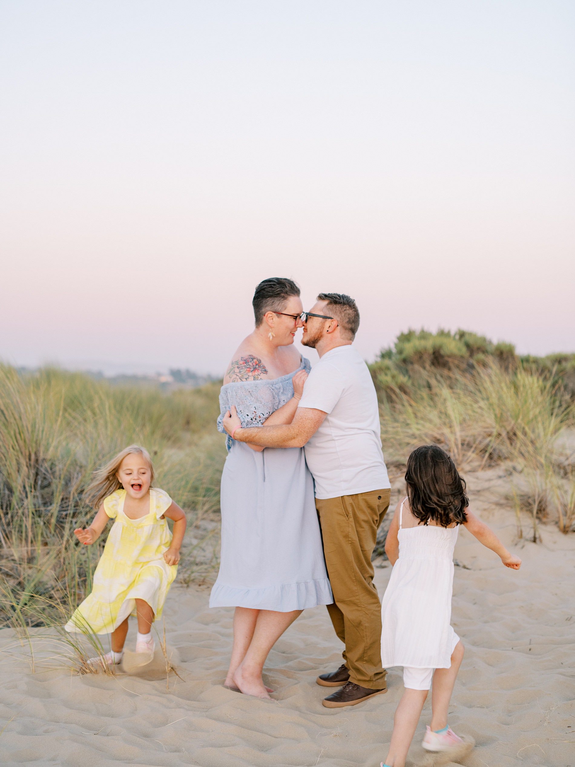 Mom and dad hold each other as their two daughters run around them in a circle agains the pink and blue sunset sky at the Oceano Dunes by Pismo Beach Family Photographer Austyn Elizabeth Photography