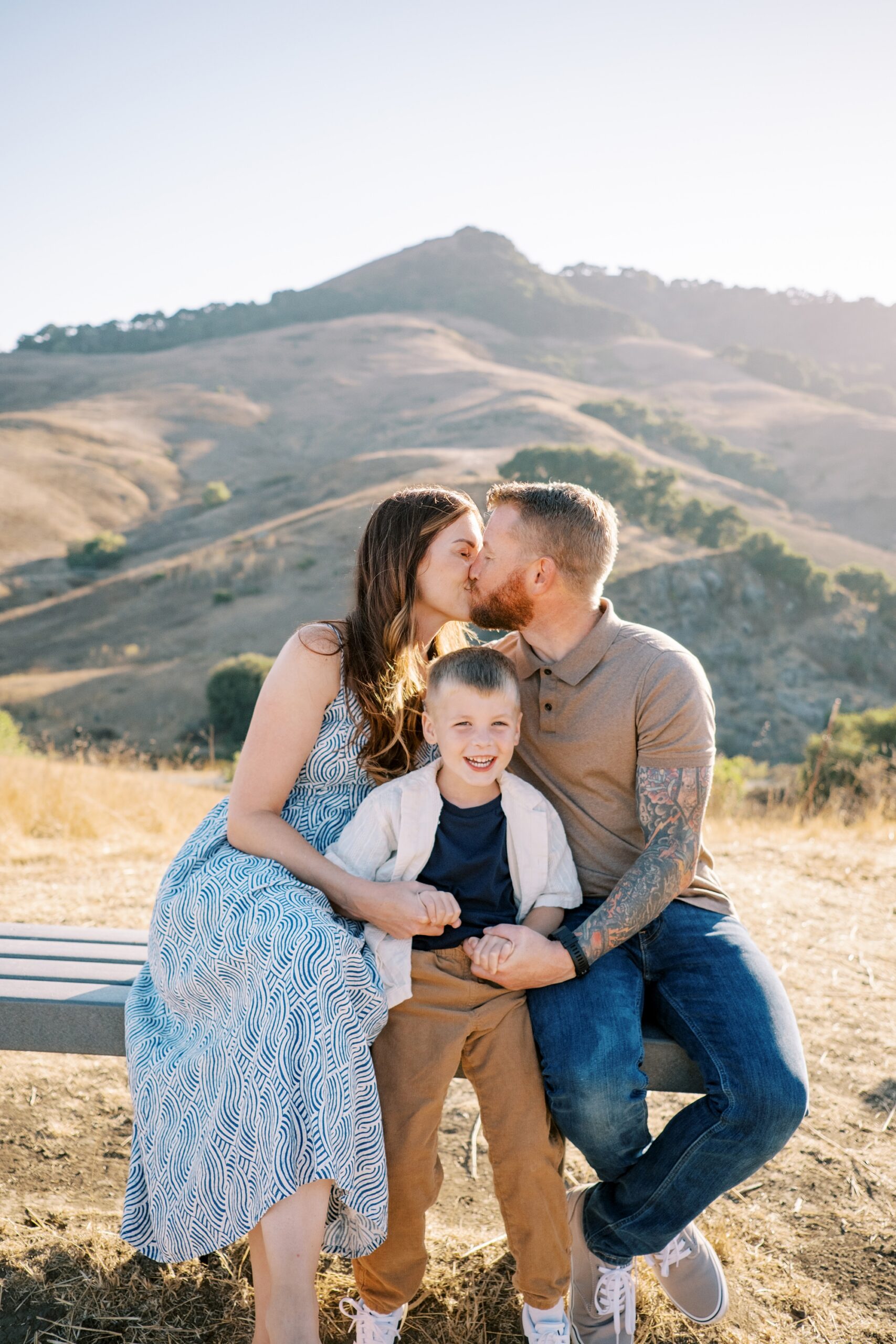 Mom and dad kiss while little boy smilies at the camera at Johnson Ranch Trail by Pismo Beach Family Photographer Austyn Elizabeth Photography