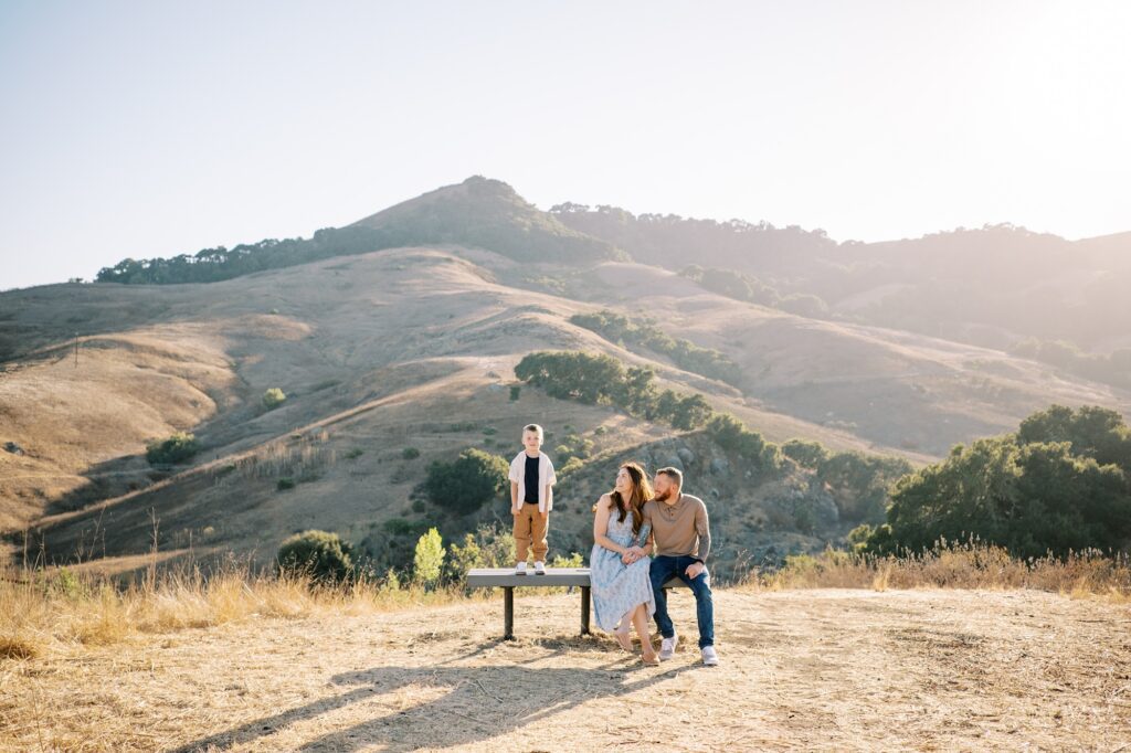 Family of three sitting on bench with SLO hills in the background at Johnson Ranch Trail by Pismo Beach Family Photographer Austyn Elizabeth Photography