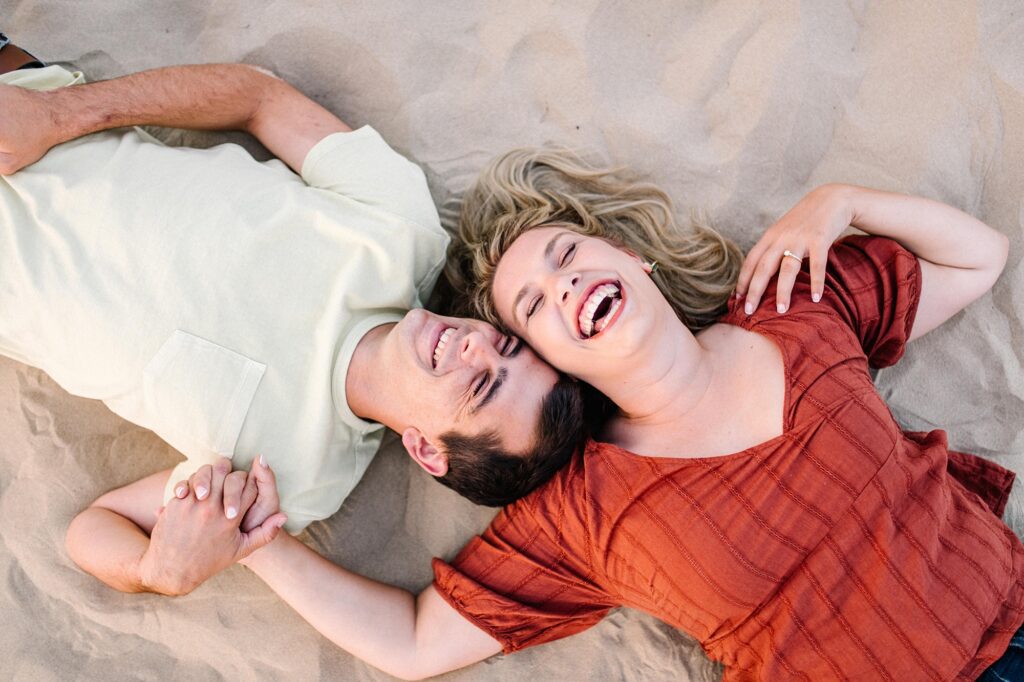 Couple laugh while laying in the sand dunes in pismo beach by Pismo Beach Engagement Photographer Austyn Elizabeth Photography