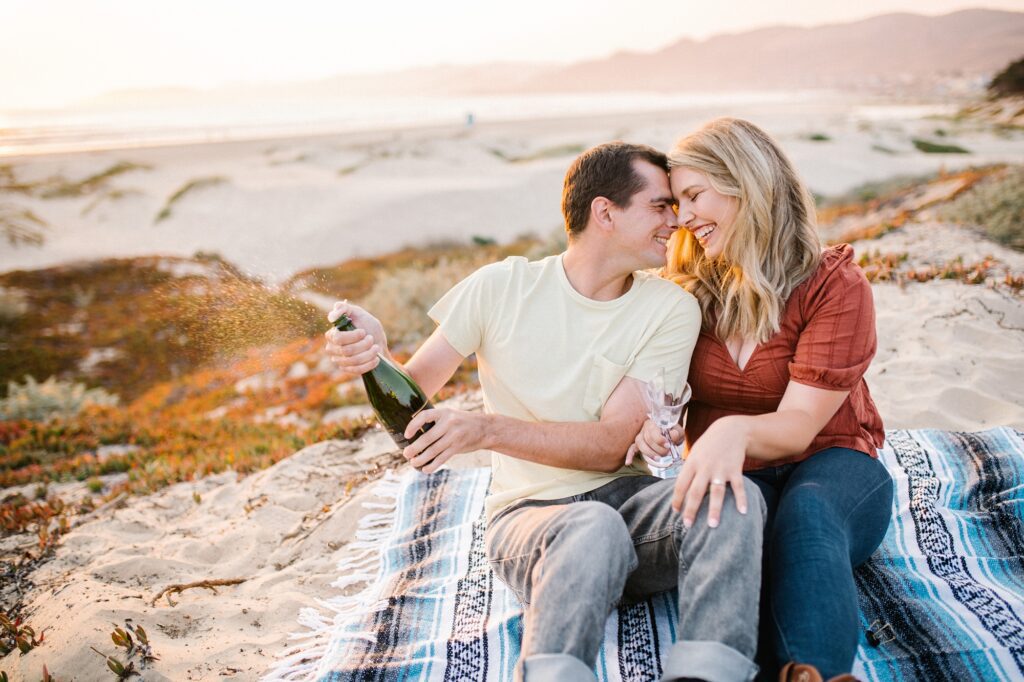 Engaged couple open champagne on beach blanket with ocean in the background during engagement session by Pismo Beach Engagement Photographer Austyn Elizabeth Photography