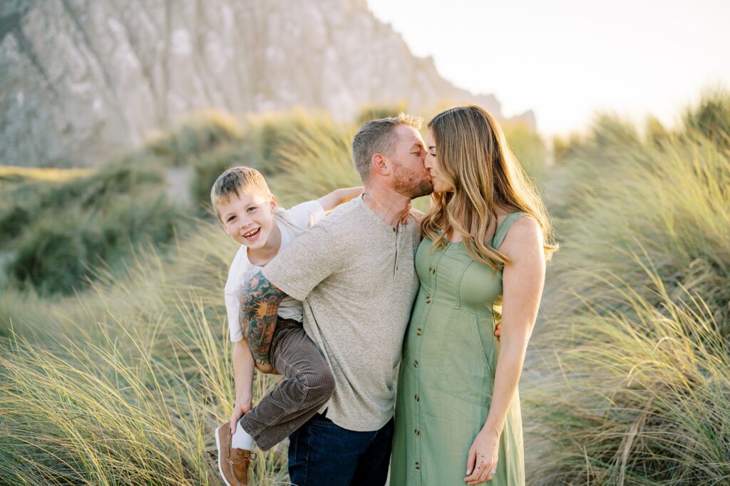Mom and dad kiss with eight year old getting piggy back ride near morro rock by Morro Bay Family Photographer Austyn Elizabeth Photography